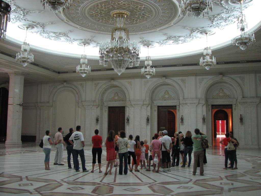 A distance shot of a room with a hanging chandelier also hanging form the ceiling which is decorated with circular decorative mouldings. 