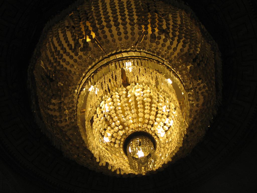 A large chandelier with a round bulbous top piece. The smaller and lighter round lower piece shines with a golden light.
