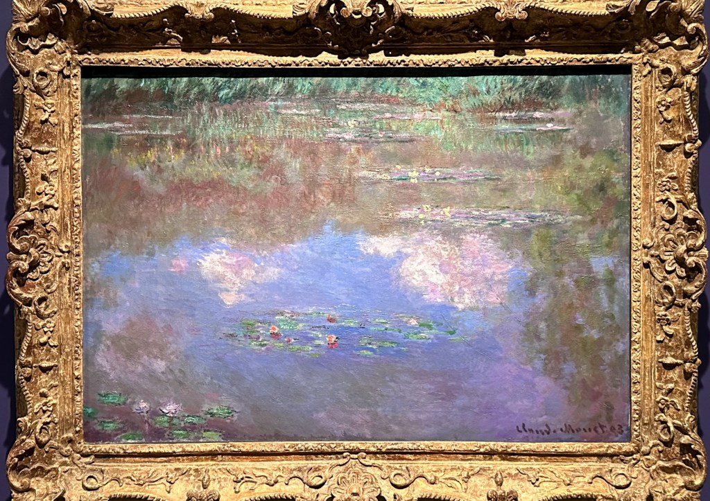 Photo of white clouds and green trees reflected in the blue of the water. A group of water lilies floats in the centre of the picture with two more distant groups further back to the right and a few even more distant groups towards the back of the canvas. There is a part of a group in the front left hand corner. This picture shows the heavy gold frame.