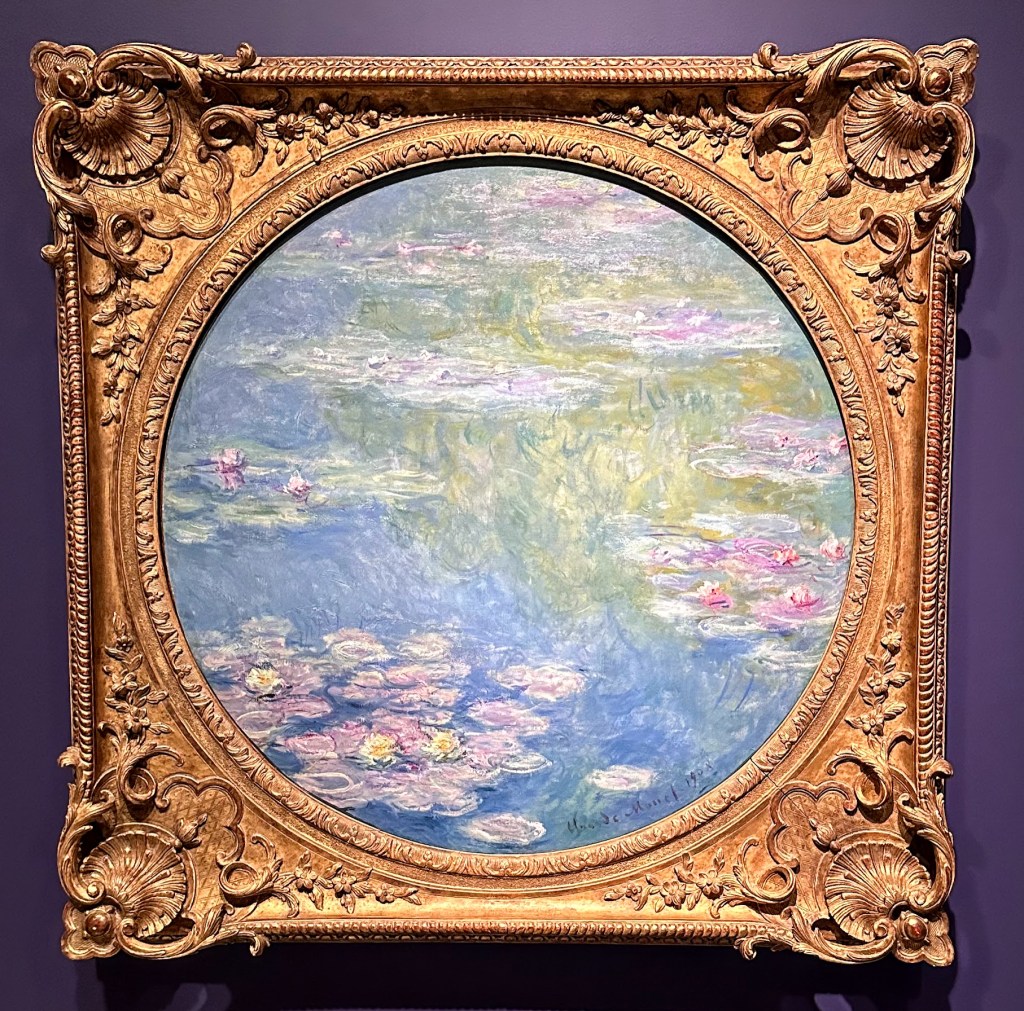 Picture caption: This is a round Water Lilies painting depicting green trees and scattered white clouds reflected int he water. The painted water lilies are much closer and you can see the individual petals in yellow and pink. The heavy gold frame surrounds the painting.